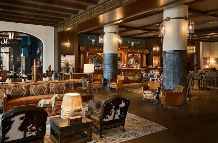 A stylish hotel lobby featuring a mix of rustic and modern furniture, including leather chairs and a decorative sofa. A wooden bar is visible in the background, illuminated by warm lighting. The space is adorned with artwork and features large columns, creating an inviting atmosphere.