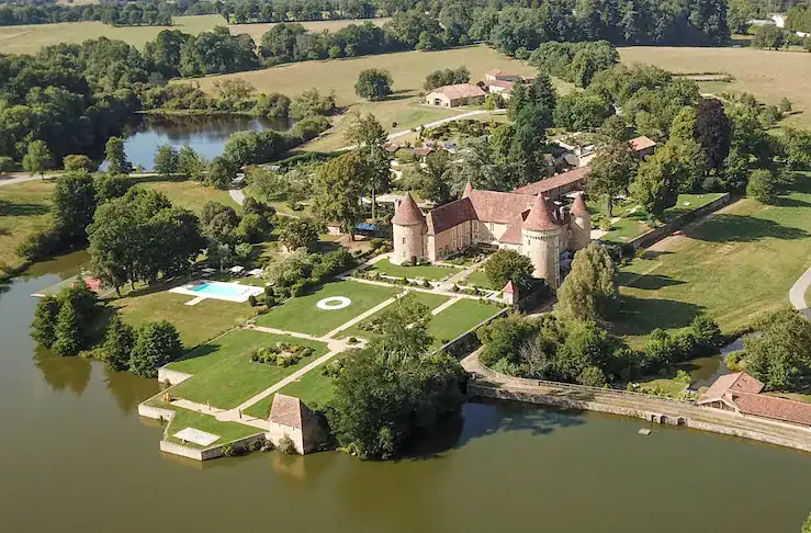 Aerial view of a picturesque estate featuring a large stone castle with turrets, surrounded by lush greenery, gardens, and a swimming pool, adjacent to a tranquil lake.