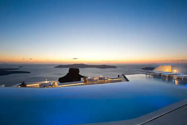 A serene infinity pool overlooks the calm sea and islands at sunset, with soft orange and blue hues in the sky. Elegant outdoor seating surrounds the pool, creating a tranquil atmosphere.