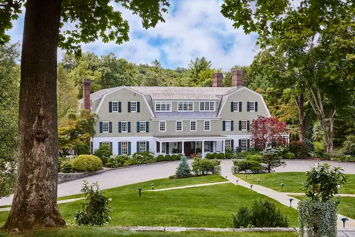 A large, elegant green and white house with multiple windows and a sloping roof, surrounded by neatly manicured lawns, trees, and shrubs, set against a backdrop of rolling hills and a cloudy sky.