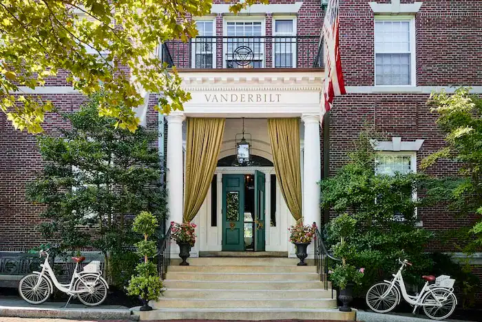 Facade of the Vanderbilt building featuring large entry steps, green doors with draped curtains, and a decorative lantern; flanked by potted plants and two vintage bicycles.