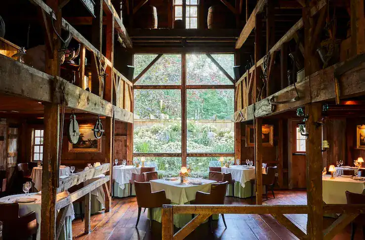 Interior of a rustic restaurant featuring wooden beams, tables set with white tablecloths and elegant dining ware, and large windows offering a view of a garden outside.