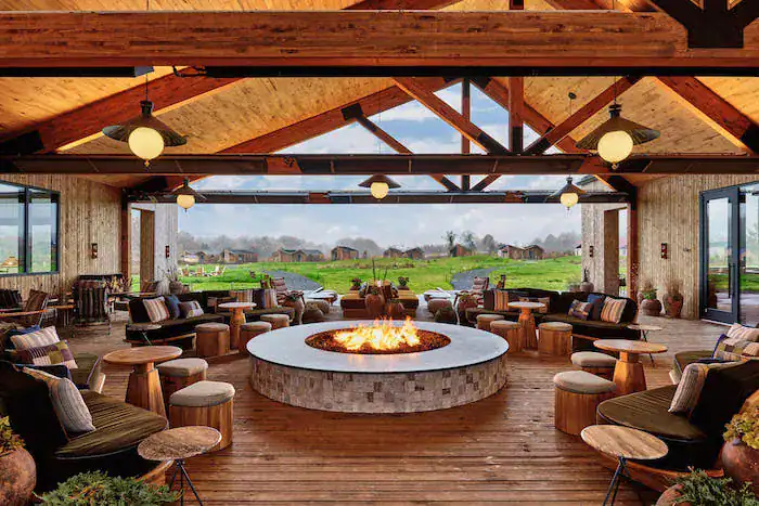A spacious, modern gathering area with a high wooden ceiling, featuring a central circular fire pit surrounded by comfortable seating. Large windows offer views of the green landscape beyond, with rustic decor and stylish furniture enhancing the inviting atmosphere.
