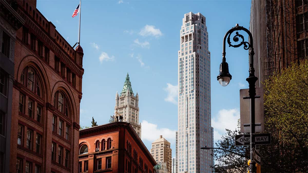 A cityscape featuring a mix of architectural styles, including a red brick building at the forefront, a green-capped skyscraper in the middle, and a modern tall building in the background against a blue sky with clouds. A streetlamp and a "One Way" sign are visible on the right.