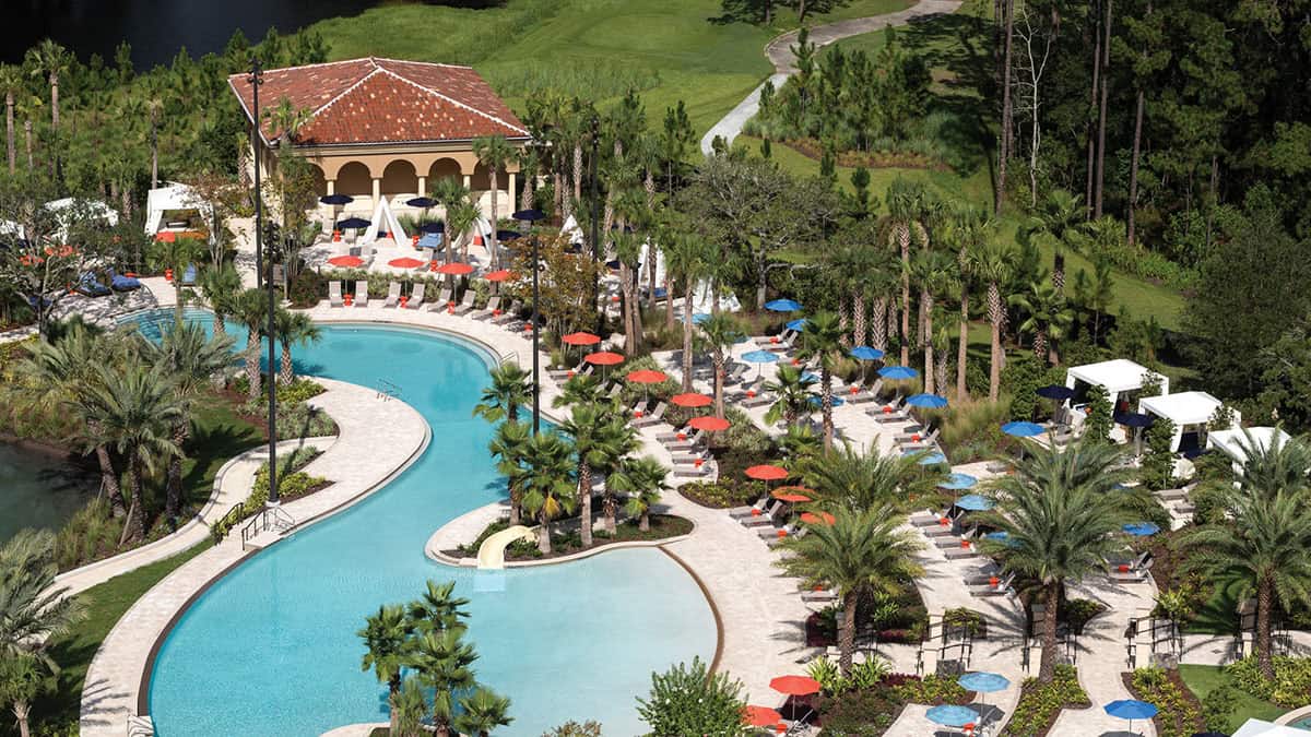 Aerial view of a resort-style pool featuring a winding shape, surrounded by palm trees and lounge chairs with colorful umbrellas. In the background, there's a building with a red tiled roof and green landscaping leading to a golf course.
