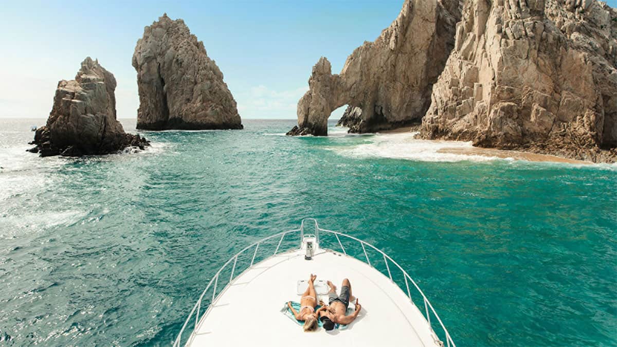 A couple relaxes on the bow of a boat, surrounded by turquoise waters and rugged rock formations under a clear blue sky.