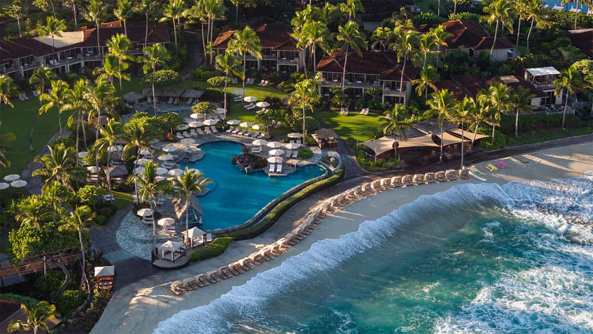 Aerial view of a tropical resort featuring a winding pool surrounded by palm trees and lounge chairs, with beachfront access and gentle waves lapping at the shore.
