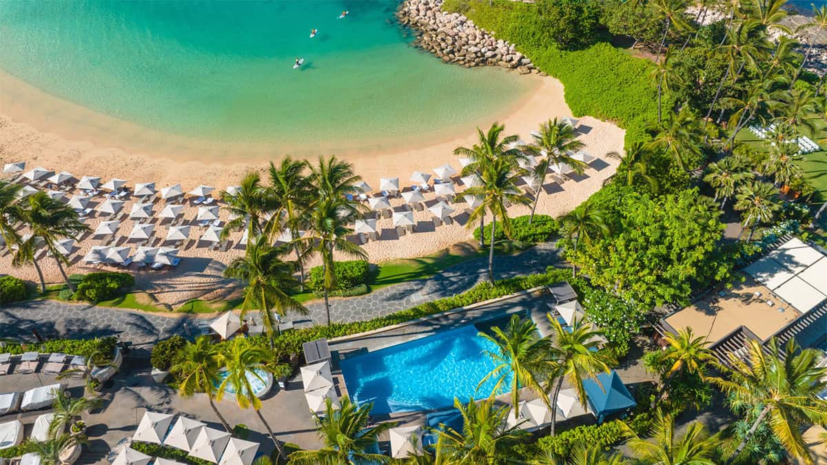 Aerial view of a tropical beach with turquoise water, white sand, and rows of umbrellas. A swimming pool with palm trees is visible beside the beach, along with lounge chairs and lush greenery in the surrounding area.