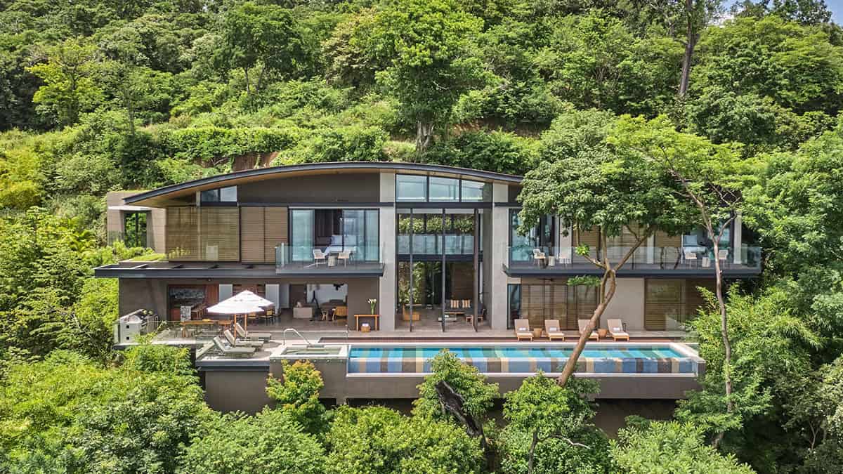 A modern two-story house nestled in lush greenery, featuring large windows, a swimming pool, and outdoor seating areas. The design includes curved rooflines and is surrounded by dense trees, offering a serene and private atmosphere.