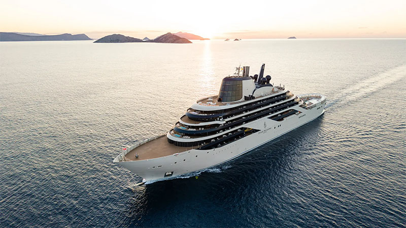 A luxury yacht sails through calm waters, with a sunset illuminating the horizon and silhouetting distant islands in the background.