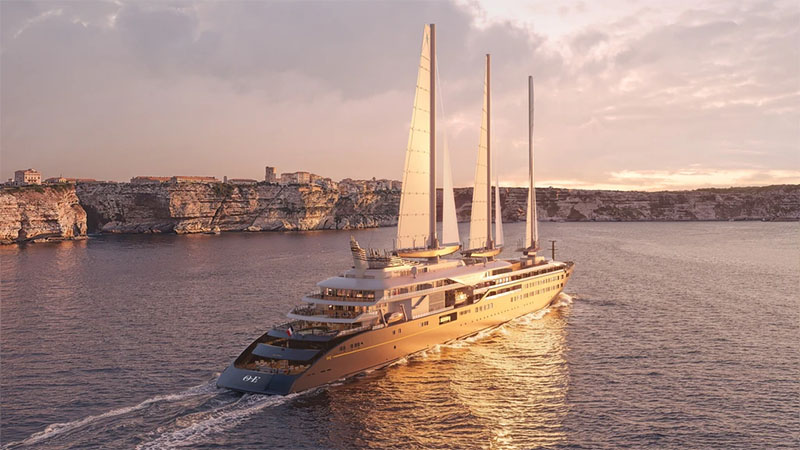 Orient Express Sailing Yachts A luxurious sailing yacht with three tall masts glides across calm waters during sunset, with rocky cliffs and a distant town visible in the background.