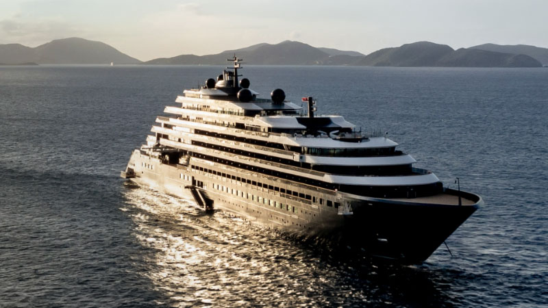 The Ritz-Carlton Yacht Collection A luxurious yacht with multiple decks is navigating through calm waters, set against a backdrop of distant mountains and a golden sunset.